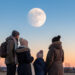 Un homme, une femme, un jeune garçon et une jeune fille emmitouflés observent une Lune partiellement éclipsée au-dessus d’un horizon ouest dégagé à l’aube.