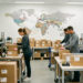 Entrepôt e-commerce moderne où une femme et deux hommes préparent des colis internationaux, avec une carte du monde abstraite en arrière-plan.